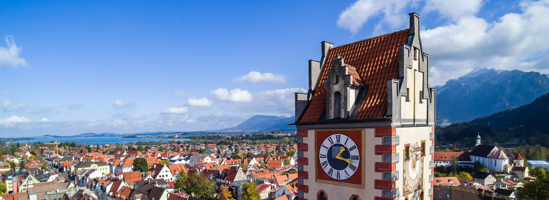 Das Foto zeigt Füssen von oben. Den Blick des Betrachters wird über die Dächer von Füssen geleitet. Im Vordergrund erhebt sich der sogenannte Uhrturm des Hohen Schlosses. Dahinter beginnen die Dächer der Füssener Altstadt. Im Hintergrund kann man schon die Berge und den türkisblauen Forggensee erkennen.