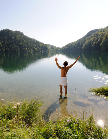 Das Bild zeigt einen Mann, der sich nur ein weißes Handtuch um die Hüften gebunden hat. Er steht im knöcheltiefen Wasser eines Sees und streckt die Arme nach oben.