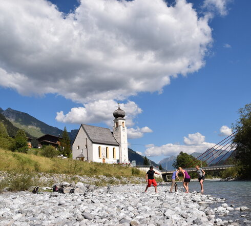 Das Bild zeigt vier Personen, die in einem Bachbett stehen. Das Ufer es Bachbetts ist mit groben Steinen und Kies bedeckt. Sie haben die Schuhe ausgezogen und treten ins Wassern. Im Hintergrund steht eine Kirche mit Kirchturm. Dahinter erheben sich die Berge. Der Himmel ist blau. Die Sonne scheint.
