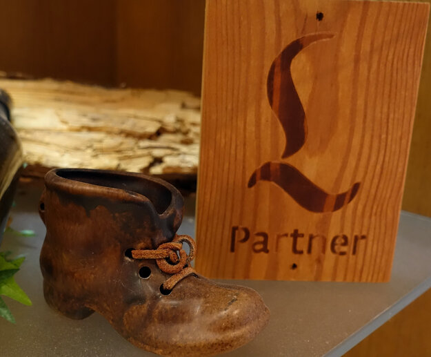 Das Bild zeigt ein Holzschild, auf dem ein großes „L“ eingebrannt ist. Darunter ist „Partner“ zu lesen. Daneben steht ein Model eines Bergschuhs aus Metall und eine Bierflasche.  