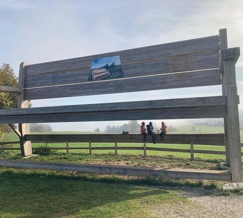 Auf dem Bild ist eine überdimensional große Sitzbank aus Holz zusehen, die in einer nebenligen ländlichen Landschaft steht. Am Fuß der Bank sitzen 3 Kinder. 