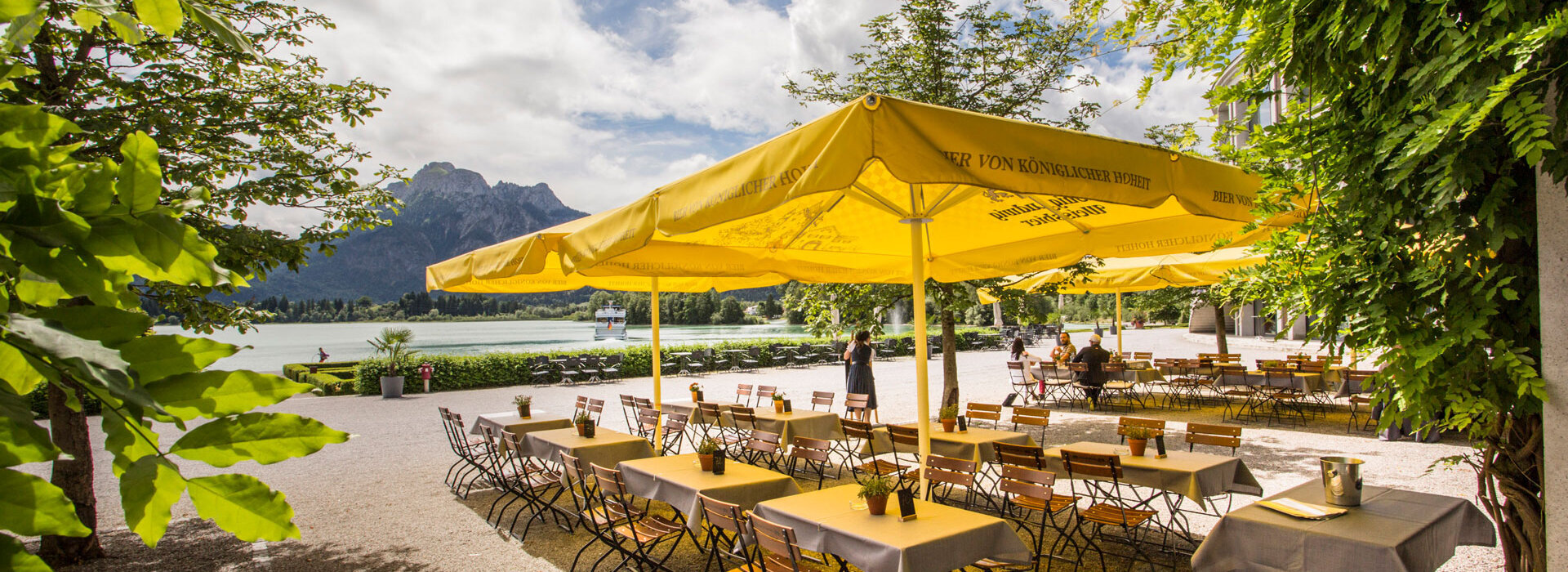 Das Foto zeigt einen Biergarten. Auf einem Kiesboden stehen Tische mit je vier Stühlen unter großen, gelben, viereckigen Sonnenschirmen. Vier Personen sitzen an den hinteren Tischen und unterhalten sich. Der Blick wird über die Tisch zum Forggensee und den dahinter liegenden Bergen gelenkt. Auf dem Forggensee fährt ein Schiff. Der Himmel ist blau mit weißen Wolken. Die Sonne scheint.