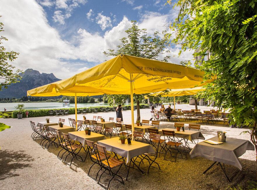 Das Foto zeigt einen Biergarten. Auf einem Kiesboden stehen Tische mit je vier Stühlen unter großen, gelben, viereckigen Sonnenschirmen. Vier Personen sitzen an den hinteren Tischen und unterhalten sich. Der Blick wird über die Tisch zum Forggensee und den dahinter liegenden Bergen gelenkt. Auf dem Forggensee fährt ein Schiff. Der Himmel ist blau mit weißen Wolken. Die Sonne scheint.