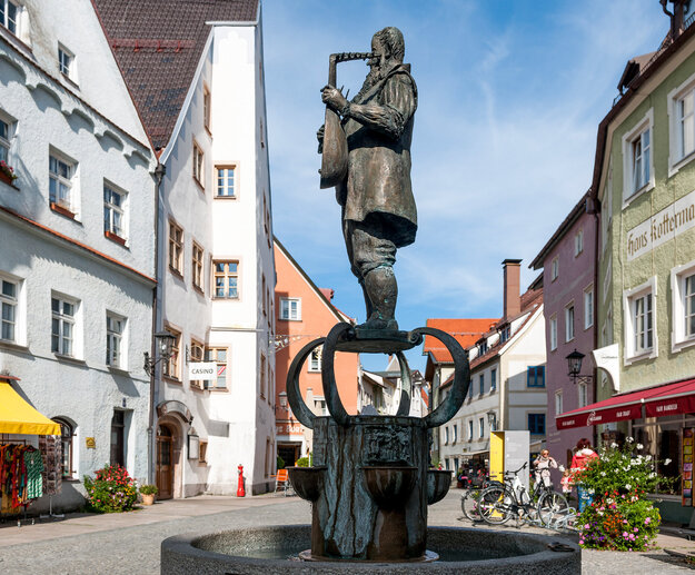 Das Foto zeigt den Lautenmacherbrunnen auf dem Brotmarkt in Füssen. In dem steinernen, runden Brunnenbassin steht ein bronzener Sockel auf dem die Bronzefigur eines bärtigen Mannes zu sehen ist, der auf einer Laute spielt. Bei der Figur handelt es sich um Caspar Tieffenbrucker einen bekannten Füssener Lautenmacher. Der Platz ist gesäumt von bunten Gebäuden. Der Himmel ist blau. Es ist Sommer.