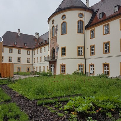 Auf dem Bild ist das Kloster Ettal zu sehen. Im Vordergrund erkennt man einen schön angelegten Kräutergarten mit grünen Planzen. 