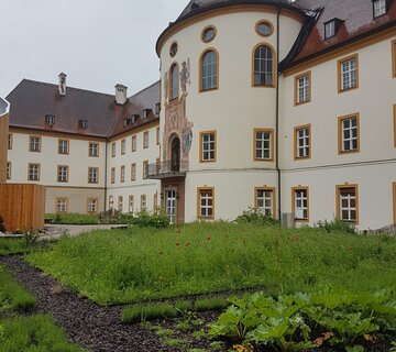 Auf dem Bild ist das Kloster Ettal zu sehen. Im Vordergrund erkennt man einen schön angelegten Kräutergarten mit grünen Planzen. 