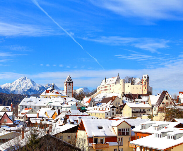 Das Bild zeigt die verschneite Füssener Altstadt mit dem imposanten Hohen Schloss im Hintergrund. Ein blauer Himmel mit Wolken und Flugstreifen bildet die Kulisse.