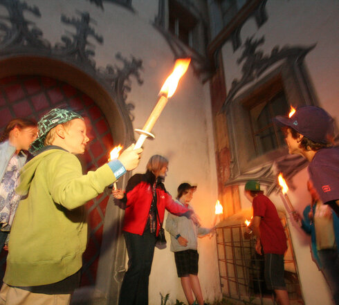 Das Bild zeigt eine lebendige Szene einer Gruppe von Menschen, die sich vor einem Gebäude versammeln. Das Gebäude, gekennzeichnet durch einen großen Bogen, dient als Kulisse für die Gruppe. Die Menschen, ein Gemisch aus Erwachsenen und Kindern, sind mit dem Aktes des Kerzenanzündens beschäftigt. Die Kerzen, von der Gruppe erhoben, brennen, und werfen ein warmes Schein auf die Szene. Die Gruppe ist vielfältig, mit Individuen unterschiedlichen Alters und Geschlechts. Einige von ihnen halten Fackeln, was zur feierlichen Stimmung beiträgt. Das Gesamtbild ist eine Feier und Einheit.