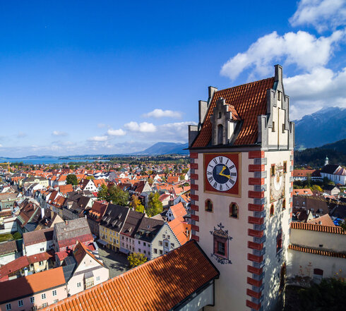 Das Foto zeigt Füssen von oben. Den Blick des Betrachters wird über die Dächer von Füssen geleitet. Im Vordergrund erhebt sich der sogenannte Uhrturm des Hohen Schlosses. Dahinter beginnen die Dächer der Füssener Altstadt. Im Hintergrund kann man schon die Berge und den türkisblauen Forggensee erkennen.