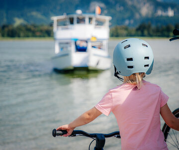 Das Bild zeigt ein Mädchen mit rosa T-Shirt und Helm, das den Lenker seines Fahrrads in der einen Hand hält. An der anderen Hand hält sie die Hand ihres Vaters. Man sieht das Mädchen nur von hinten. Sie stehen am Ufer eines Sees. Ein Schiff fährt auf den Betrachter zu. Im Hintergrund erheben sich Berge und grün, bewaldete Hügel. Der Himmel ist blau. Die Sonne scheint. 