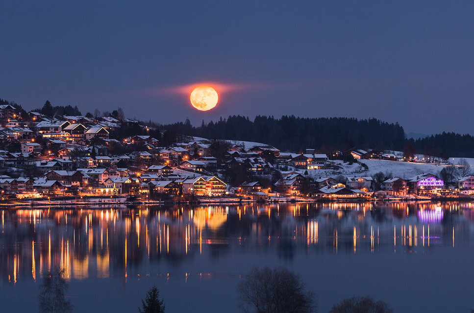 Das Bild fängt eine ruhige, winterliche Nachtszene im Ort Hopfen ein, der am Ufer des Hopfensees liegt. Der Ort wird von dem weichen Licht der Straßen- und Häuserbeleuchtung in warmes Licht getaucht, das sich im ruhigen Wasser des Sees spiegelt. In der Ferne erheben sich majestätische, schneebedeckte Berge gegen den dunklen Himmel. Der Nachthimmel wird vom Vollmond erhellt, der über die ganze Szene strahlt.