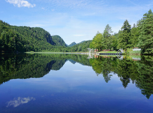 Das Bild zeigt eine ruhige Seenlandschaft. Der Obersee, der der zentrale Fokus des Bildes ist, ist von üppigem Grün umgeben und von Bergen gesäumt. Die ruhige Wasseroberfläche des Sees spiegelt die Umgebung wider. Auf der rechten Seite des Bildes befindet sich ein Steg mit einem Sprungturm. Der Himmel darüber ist klar und blau.