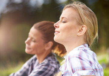 Das Bild zeigt zwei junge Frauen, die mit geschlossenen Augen das Gesicht in die Sonne halten und die Situation sichtlich genießen. Man sieht lediglich die Gesichter und einen Teil des Oberkörpers beider Frauen. Sie tragen karierete Oberteile und haben die Haare zusammengebunden. 