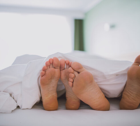 Das Bild zeigt zwei nackte Fußpaare, die unter einer weißen Decke hervorschauen. Das Bild suggeriert einen Moment der Entspannung und des Wohlbehagens.