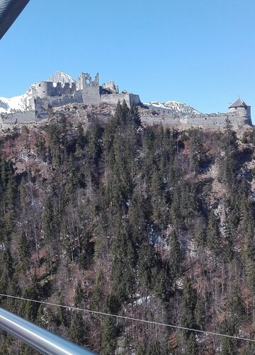 Das Bild zeigt einen atemberaubenden Ausblick auf eine Burg, die auf einem Felsen thront. Die aus Stein errichtete Burg steht majestätisch vor dem Hintergrund eines klaren blauen Himmels. Der Felsen, der mit üppigem Grün bedeckt ist, fügt der Szene die Schönheit der Natur hinzu. Eine Brücke aus Metall spannt sich über das Bild und bietet dem Betrachter eine Aussichtspunkt. Die Brücke, mit ihrer soliden Struktur, scheint ein beliebter Ort für Touristen zu sein, wie die Menschen, die zu sehen sind, die über sie gehen. Die gesamte Szene ist ein harmonisches Zusammenspiel von Architektur und Natur und bietet eine einzigartige Perspektive auf die Burg und ihre Umgebung.