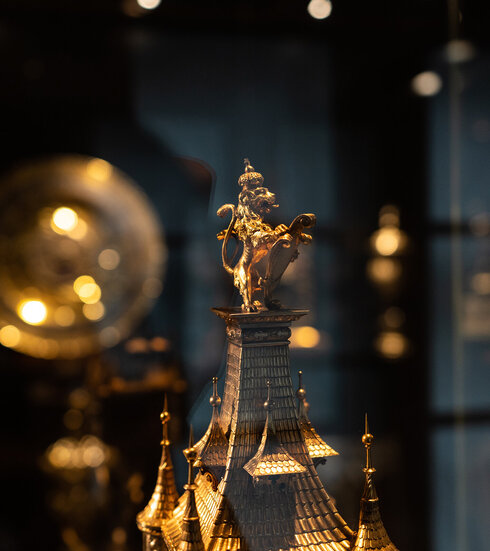 Im Fokus des Bildes ist ein goldener Löwe zu sehen, der eine Krone trägt und ein Schild hält. Der Löwe steht auf dem Dach eines Turmes. Alles ist aus Gold. Bei dem Stück handelt es sich möglicherweise um eine Ausstellungsstück in einem Museum. Im Hintergrund sind weitere goldene Gegenstände sichtbar doch nur schwer zu erkennen. 