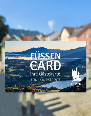 Das Bild zeigt eine Karte, in der Größe einer Scheckkarte. Diese ist mit einem Panoramafoto bedruckt und trägt die Aufschrift „Füssen Card, Ihre Gästekarte, Your guestcard“ Im Hintergrund sieht man unscharf bunte Häuserfassaden. Der Himmel ist blau. Die Sonne scheint.