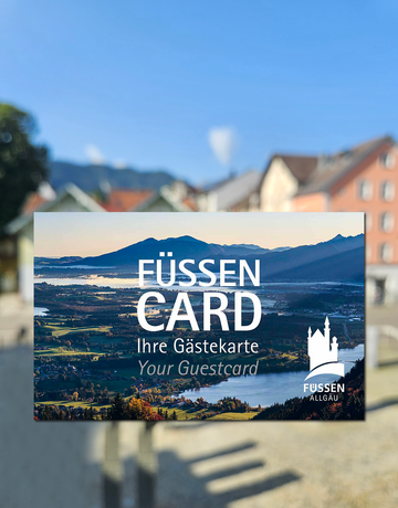 Das Bild zeigt eine Karte, in der Größe einer Scheckkarte. Diese ist mit einem Panoramafoto bedruckt und trägt die Aufschrift „Füssen Card, Ihre Gästekarte, Your guestcard“ Im Hintergrund sieht man unscharf bunte Häuserfassaden. Der Himmel ist blau. Die Sonne scheint.