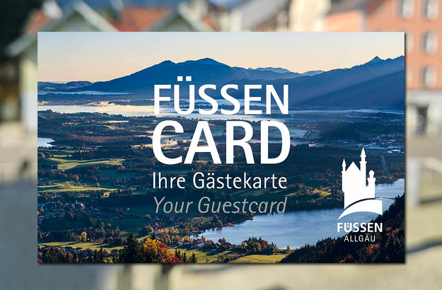 Das Bild zeigt eine Karte, in der Größe einer Scheckkarte. Diese ist mit einem Panoramafoto bedruckt und trägt die Aufschrift „Füssen Card, Ihre Gästekarte, Your guestcard“ Im Hintergrund sieht man unscharf bunte Häuserfassaden. Der Himmel ist blau. Die Sonne scheint.