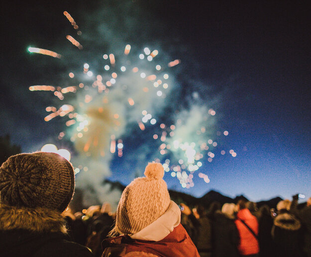 Das Bild zeigte ein Feuerwerk am dunklen Nachthimmel. Zwei Personen stehe im Vordergrund. Nur ihre Köpfe und Schultern sind zu sehen. Sie tragen Winterkleidung und Mützen und sind vom Betrachter weggewandt.  