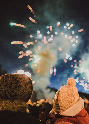 Das Bild zeigte ein Feuerwerk am dunklen Nachthimmel. Zwei Personen stehe im Vordergrund. Nur ihre Köpfe und Schultern sind zu sehen. Sie tragen Winterkleidung und Mützen und sind vom Betrachter weggewandt.  