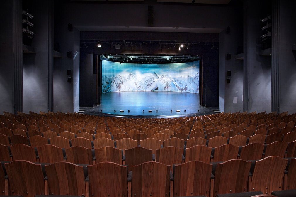 Das Foto zeigt den Saal des Festspielhauses Neuschwanstein. Der Blick geht über schier unzählige, braune Holzstuhllehnen hinweg zur Bühne. Das Bühnenbild zeigt schneebedeckte Berge. Der Zuschauerraum ist menschenleer. 