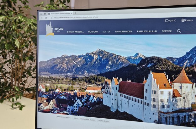 Das Bild zeigt einen Teil eines Bildschirms. Dort ist die Website von Füssen zu sehen. Der Bildschirm ist fast vollständig mit einem Foto ausgefüllt, das das Hohe Schloss zeigt, dass über eine Anhöhe über Füssen thront. Im Hintergrund des Bildes erheben sich die teilweise schneebedeckten Berge. Hinter dem Bildschirm ist eine Zimmerpflanze zu sehen. 