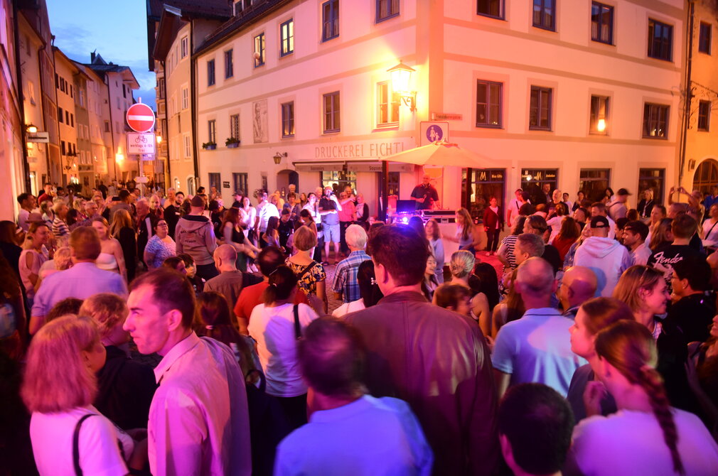 Das Bild zeigte vier eine Kreuzung in der Altstadt. Die Kreuzung und auch die beiden weiterführenden Gassen sind mit Menschen gefüllt. In der Mitte vor einem Hauseck steht ein DJ-Pult. Gerade geht die Sonne unter. Die Straßenbeleuchtung ist schon an und taucht die Szenerie in warmes Licht. Die Menschen tanzen und reden miteinander. Die Stimmung wirkt locker und fröhlich. 