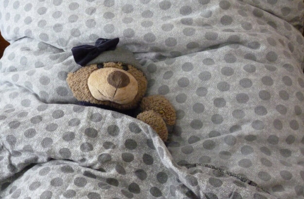 Das Bild zeigt ein graues Kissen und eine graue Bettdecke. In das Bett wurde ein Teddybär gelegt. Er trägt eine graue Mütze mit einer schwarzen Schleife.   