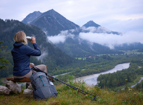 Das Bild zeigt eine Frau, die auf einem Holzscheit auf einer Anhöhe sitzt und ins Tal hinabblickt. Dort fließt ein Fluss. Sie trinkt aus einer Metallflasche und trägt Wanderkleidung. Neben ihr liegen Wanderstöcke. Im Hintergrund erheben sich bewaldete Berge. Der Himmel ist wolkenverhangen. Nebel zieht auf.