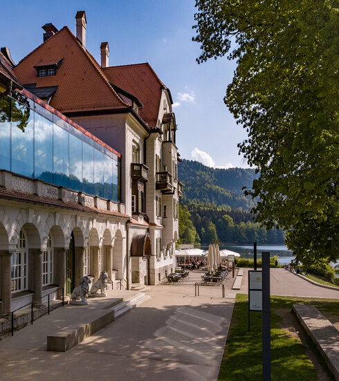 Man blickt entlang des Gebäudes des Museums der bayerischen Könige. Im Hintergrund erheben sich die Berge. Der Bergwald beginnt, davor ist der Alpsee zu sehen. Am Ufer des Alpsees sieht man die Terrasse des Restaurants Alpenrose am See. Unter Sonnenschirmen sieht man mehrere Personen an Tischen sitzen. Links und rechts neben dem grünen Portal des Museums stehen zwei lebensgroße, steinerne Löwen.  