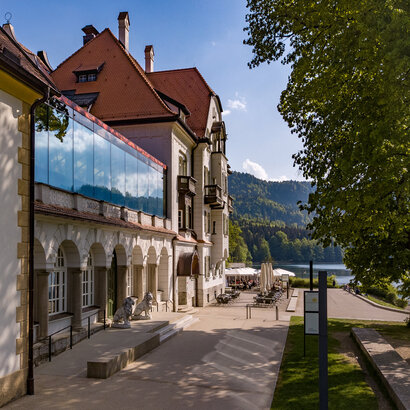 Man blickt entlang des Gebäudes des Museums der bayerischen Könige. Im Hintergrund erheben sich die Berge. Der Bergwald beginnt, davor ist der Alpsee zu sehen. Am Ufer des Alpsees sieht man die Terrasse des Restaurants Alpenrose am See. Unter Sonnenschirmen sieht man mehrere Personen an Tischen sitzen. Links und rechts neben dem grünen Portal des Museums stehen zwei lebensgroße, steinerne Löwen.  