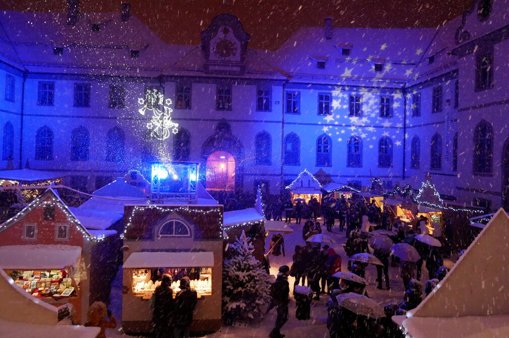 Das Bild zeigt den Füssener Adventsmarkt im Klosterhof des ehemaligen Benediktinerklosters St. Mang am Abend. Die Fassade des rundumlaufenden Gebäudes ist blau beleuchtet. Die schneebedeckten Buden des Adventsmarkt sind ebenfalls festlich beleuchtet. Zahlreiche Menschen sind zu sehen. Es schneit.   