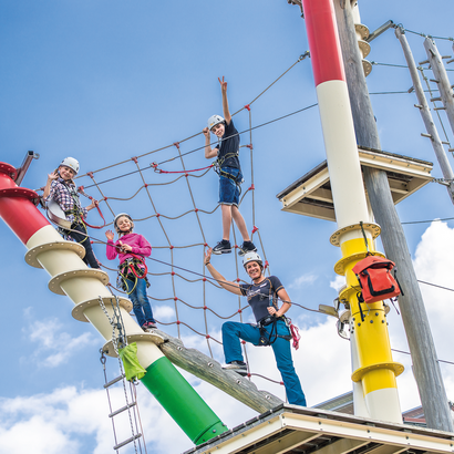 Das Foto zeigt einen Teil eines Kletterparks, dessen Stangen wie Mikado Stäbe lackiert sind. Zwischen den Seilen und Netzen, die die unterschiedlichen Stangen verbinden, sind Trittstufen und Plattformen unterschiedlicher Größe angebracht. Dort sitzen, stehen und balancieren drei Kinder und eine Frau, die in die Kamera blicken und winken. Der Himmel ist blau mit wenigen weißen Wolken durchzogen. 