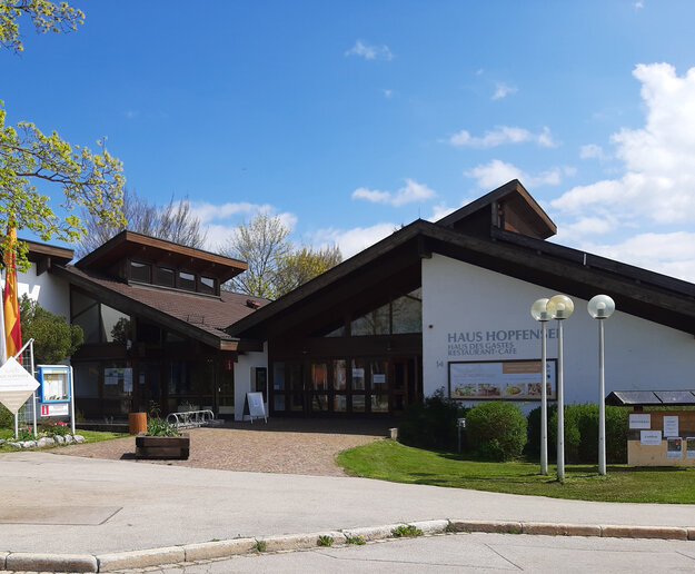 Das Bild zeigt ein die Vorderseite des Hauses Hopfensee. Das Gebäude besteht scheinbar aus zwei Häusern, die an ihren Dächern zusammengefügt wurden. Vor dem Gebäude stehen Fahnenmasten mit unterschiedlichen Fahnen und Lampen. Eine Weg führt auf den Haupteingang zu.  