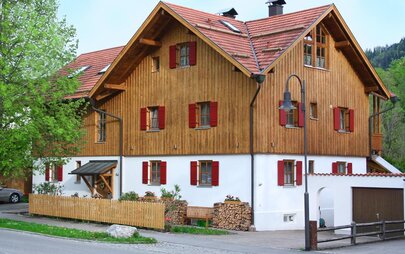 Das Bild zeigt ein zweistöckiges Haus, das ab dem 1. Stock mit Holz verkleidet ist. Die Fenster umrahmen rote Fensterläden. Im Hintergrund sind hohe Bäume zu sehen.   