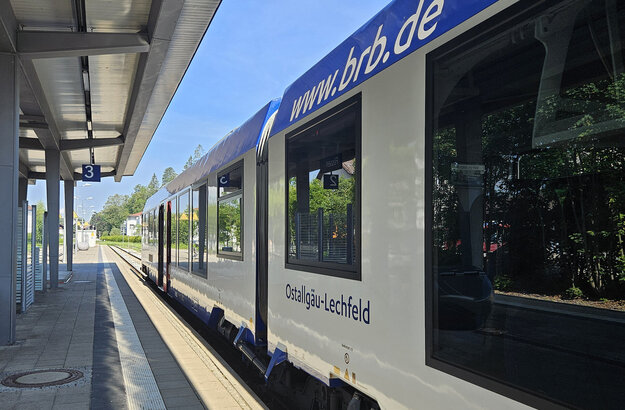 Das Bild zeigt einen Zug, der an einem Bahnsteig steht. Der Zug ist weiß-blau und trägt die Aufschrift: www.brb.de sowie „Ostallgäu-Lerchenfeld“. Der Bahnsteig trägt die Nummer 3 und ist überdacht. Der Himmel ist blau. Die Sonne scheint.