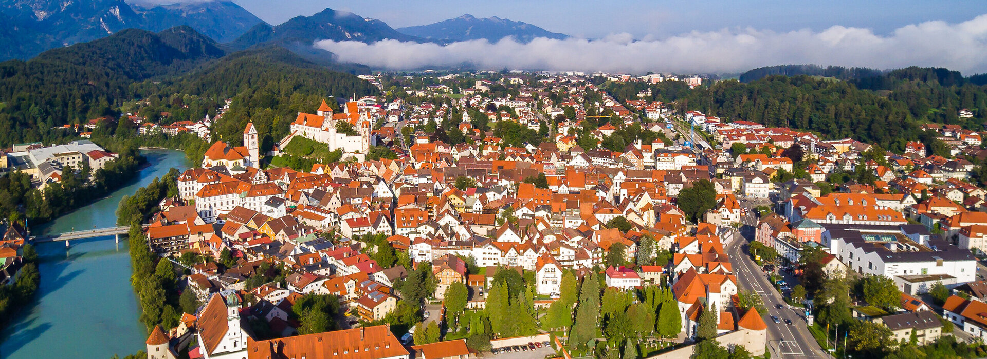 Das Foto zeigt Füssen von oben. Den Blick des Betrachters wird über die Dächer von Füssen geleitet. Im Vordergrund ist das Franziskanerkloster zu erkennen, das aus Klostergebäude und Klosterkirche St. Stephan besteht. Das Klostergelände ist eingerahmt von der historischen Stadtmauer. Neben dem Kloster fließt der türkisblau Fluss Lech. Im Hintergrund des Klosters erstreckt sich die Füssener Altstadt mit dem Hohen Schloss und dem Bergpanorama.