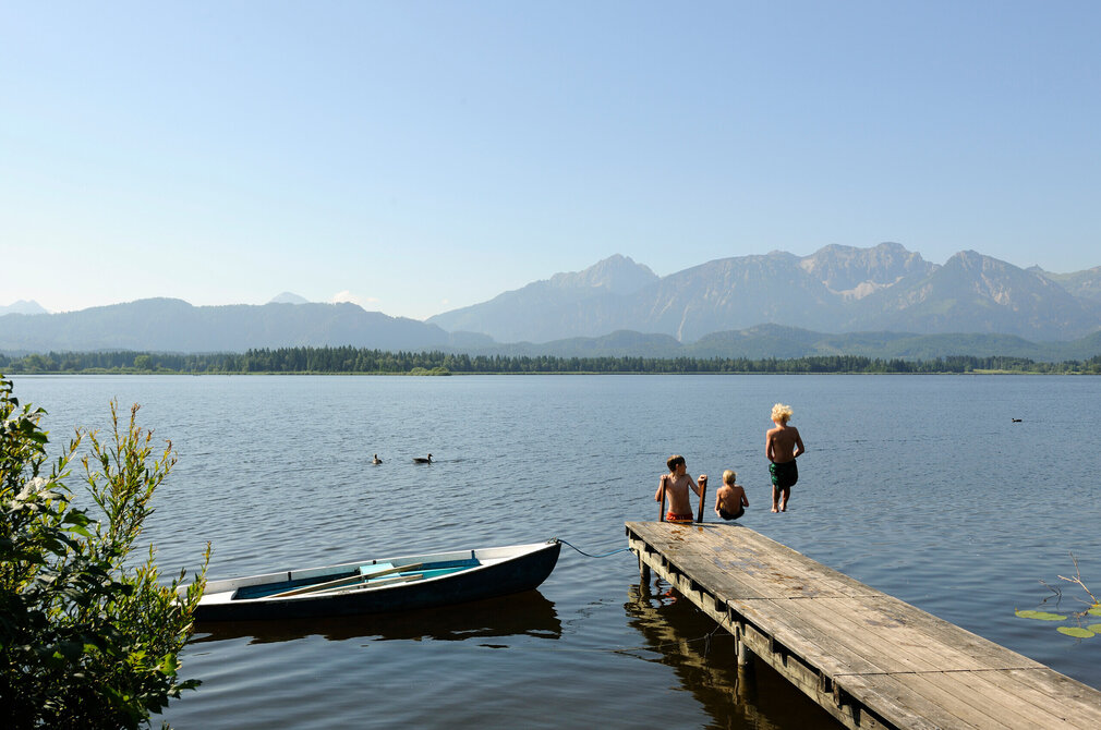 Die Aufnahme erfasst eine idyllische Szenerie am Hopfensee. Im Vordergrund erstreckt sich ein hölzerner Steg ins Wasser, an dem sich drei Kinder tummeln. Sie springen vom Steg ins Wasser oder klettern gerade die Leiter aus dem Wasser zurück auf den See. Der Steg ist auf der rechten Seite des Bildes zu sehen. Im Hintergrund erheben sich majestätisch Berge, deren Spitzen den klaren blauen Himmel berühren. Die Berge bilden eine atemberaubende Kulisse für den ruhigen See. Links vom Bild liegt ein kleines blaues Boot am Steg angedockt.