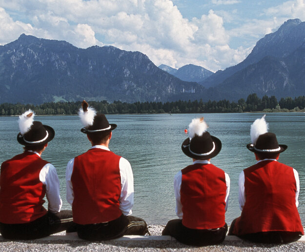 Das Bild zeigt eine ruhige Szene von vier Personen, die alle in traditioneller Kleidung gekleidet sind und am Ufer des Forggensees sitzen. Die Kleidung besteht aus roten Jacken, schwarzen Hüten, deren Bedeckung mit weißen Federn verziert sind, und weißen Hemden.