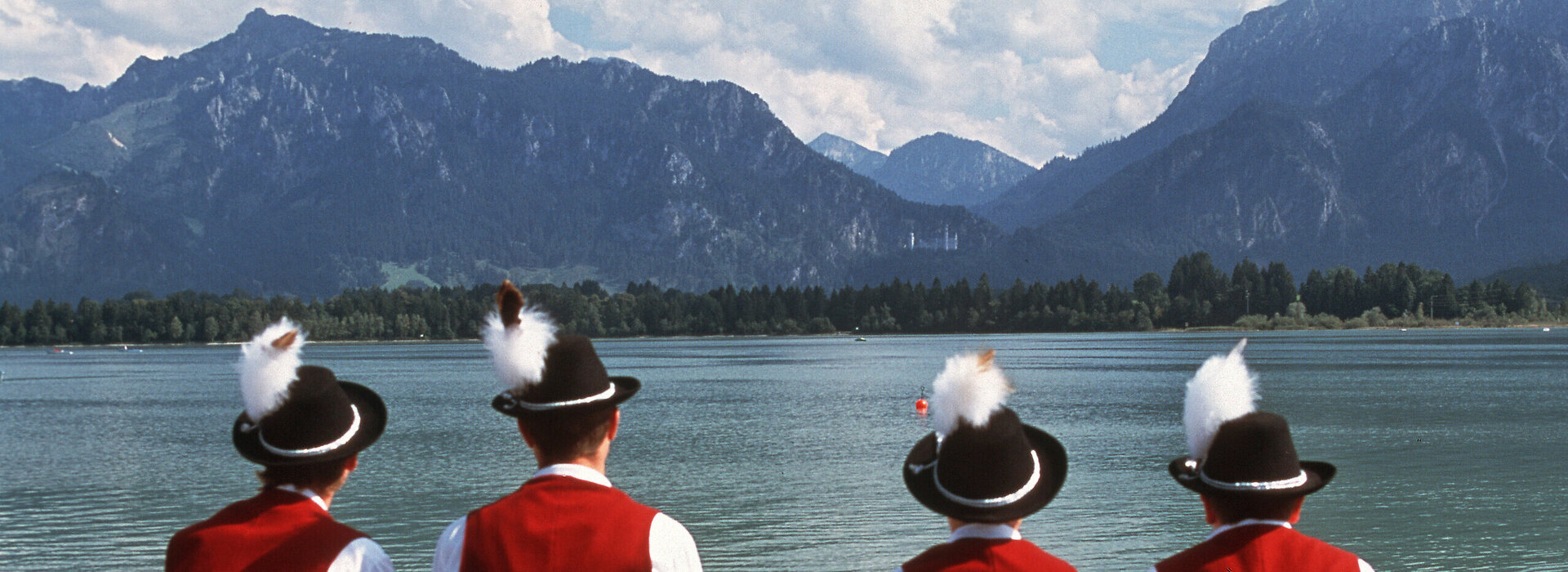 Das Bild zeigt eine ruhige Szene von vier Personen, die alle in traditioneller Kleidung gekleidet sind und am Ufer des Forggensees sitzen. Die Kleidung besteht aus roten Jacken, schwarzen Hüten, deren Bedeckung mit weißen Federn verziert sind, und weißen Hemden.