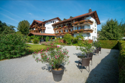 Das Bild zeigt einen mehrstöckigen Gebäudekomplex. Jedes Stockwerk hat einen rundumlaufenden Balkon, der mit blühenden Blumenkästen geschmückt ist. Das Haus ist umgeben von hohen Bäumen und Büschen. Auf dem Haus ist „Filser“ zu lesen. Ein breiter Kiesweg führt zu dem Haus. Auf dem Kiesweg stehen ebenso Blumentöpfe, Tische und Stühle. 