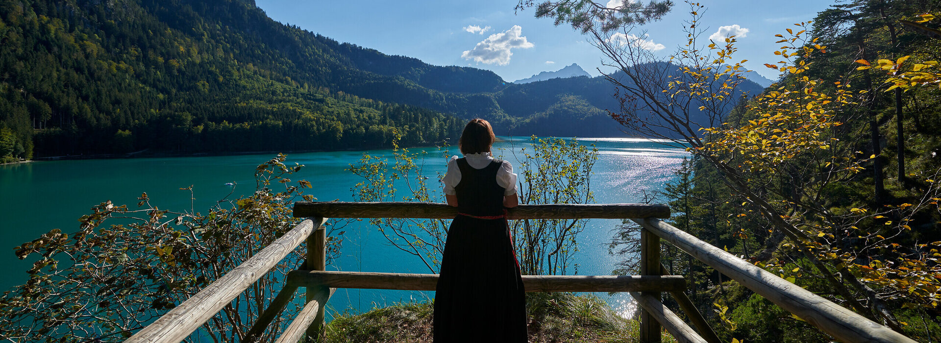 Eine Frau steht an einem Aussichtspunkt am Ufer des türkisgrünen Lechs in Füssen