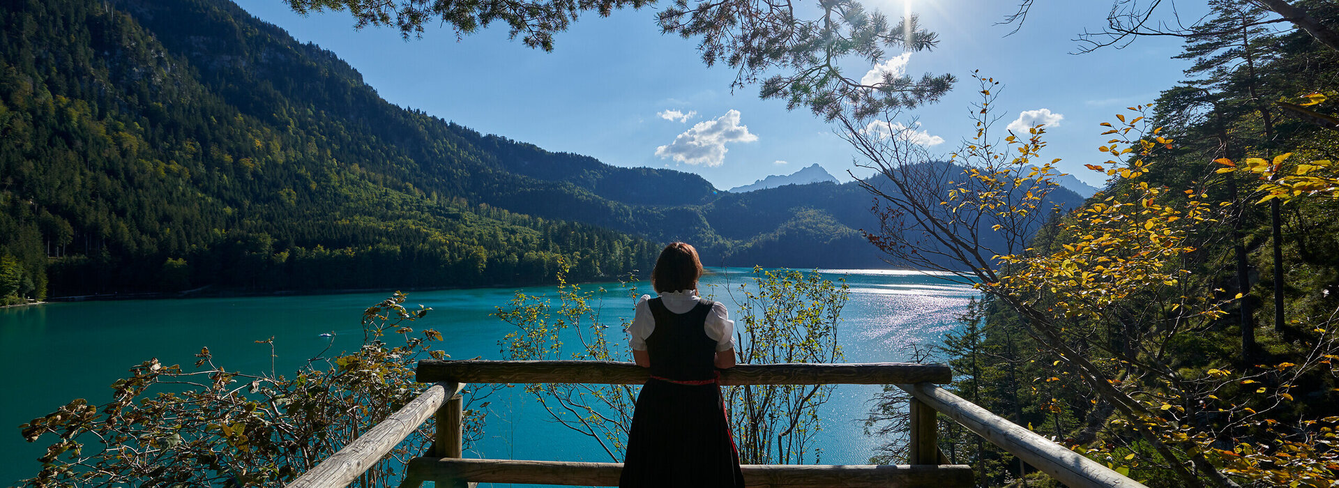 Eine Frau steht an einem Aussichtspunkt am Ufer des türkisgrünen Lechs in Füssen