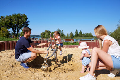 Das Bild zeigt eine herzerwärmende Szene am sandigen Strand. Ein Mann, der ein blaues Hemd und beigefarbene Shorts trägt, kniet neben einem kleinen Kind. Er hält ein metallisches Objekt, möglicherweise ein Spielzeug, in der Hand. Das Kind, das eine pinke Mütze trägt, steht neben ihm und hält sich an dem Bein des Mannes fest. Etwas weiter entfernt sitzt eine Frau auf dem sandigen Strand. Sie trägt ein weißes Hemd und blaue Shorts. Ihr Blick ist auf den Mann und das Kind gerichtet, was auf eine enge Beziehung zwischen ihnen hinweist. Der Hintergrund des Bildes zeigt eine idyllische Strandszene. Der Himmel ist klar und blau, und das Wasser erscheint ruhig. Die Gesamtstimmung des Bildes ist eine ruhige Entspannung und das Genießen eines Tages am Strand.