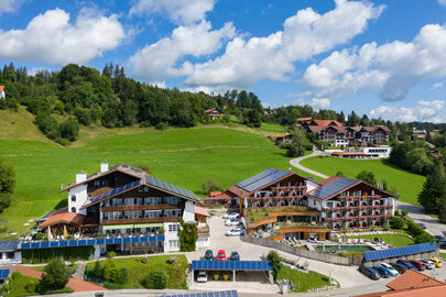 Das Bild zeigt eine malerische Ansicht eines großen Ferienresorts, das in einer Ebene angesiedelt ist. Das Resort wird gekennzeichnet durch eine Reihe von Gebäuden, jedes mit einer eigenen architektonischen Gestaltung. Die Gebäude sind hauptsächlich weiß, einige von ihnen haben rote Dächer, die dem Bild eine Farbgebung verleihen. Das Resort ist von üppigem grünen Gras umgeben, was eine friedliche und einladende Atmosphäre schafft. Eine gewundene Straße, die von Bäumen gesäumt ist, schlängelt sich durch das Resort und führt zu einem Parkplatz, wo mehrere Autos geparkt sind. Das Resort ist auf einem Hügel gelegen, was einen Panoramablick auf die umliegende Landschaft erlaubt. Der Himmel darüber ist ein klarer Blau, gesprenkelt mit flauschigen weißen Wolken, wodurch das idyllische Szenario vollendet wird.