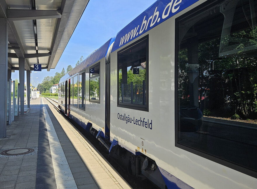 Das Bild zeigt einen Zug, der an einem Bahnsteig steht. Der Zug ist weiß-blau und trägt die Aufschrift: www.brb.de sowie „Ostallgäu-Lerchenfeld“. Der Bahnsteig trägt die Nummer 3 und ist überdacht. Der Himmel ist blau. Die Sonne scheint.