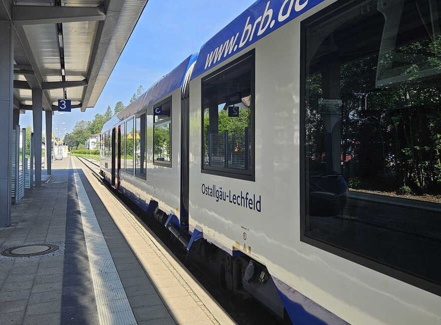 Das Bild zeigt einen Zug, der an einem Bahnsteig steht. Der Zug ist weiß-blau und trägt die Aufschrift: www.brb.de sowie „Ostallgäu-Lerchenfeld“. Der Bahnsteig trägt die Nummer 3 und ist überdacht. Der Himmel ist blau. Die Sonne scheint.