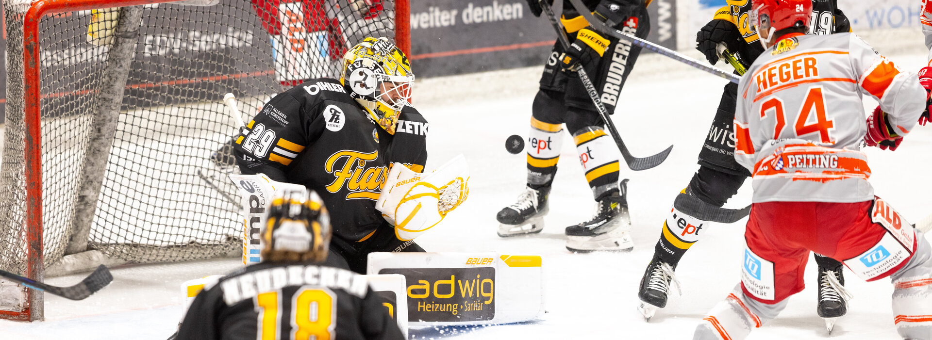 Das Bild zeigt mehrere Eishockeyspieler während eines Spiels. Sie fahren und stehe vor dem Füssener Tor. Der Torwart kniet vor dem Tor. Der Puck fliegt auf ihn zu. Alle tragen die vollständige Eishockey-Ausrüstung. Vier Spieler tragen die schwarz-gelbe Ausrüstung des E. V. Füssen. Drei die weiß-rote Ausrüstung einer Mannschaft aus Peiting. Im Hintergrund ist die Bande zu sehen.   