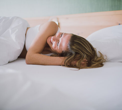 Eine Frau wacht ausgeschlafen und gut gelaunt auf, sie lächelt dabei.